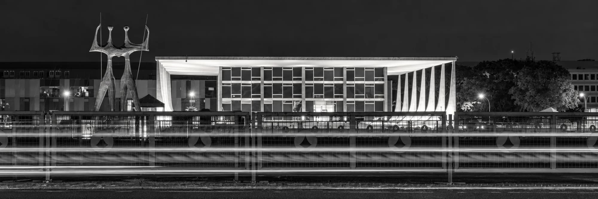 Supremo Tribunal Federal à noite, em preto e branco, com a escultura Os Candangos à esquerda e faixas de luz dos carros em primeiro plano